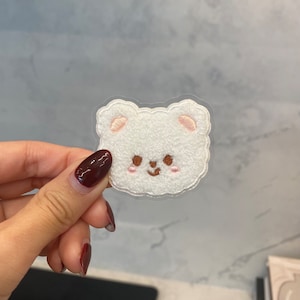 May include: White fuzzy bear face embroidered patch with pink ears and brown eyes. The patch is on a clear background.