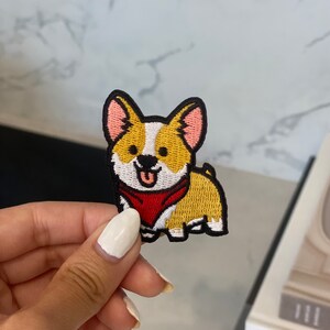 May include: Embroidered iron-on patch featuring a cartoon corgi dog wearing a red bandana. The corgi is yellow, white, and black.