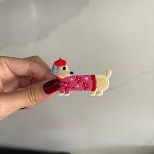 Op de afbeelding: Geborduurde teckel patch. De hond heeft een crèmekleurig lichaam, een rode baret en een roze trui met rode bloemaccenten. De patch is ontworpen om op stof te strijken of te naaien.