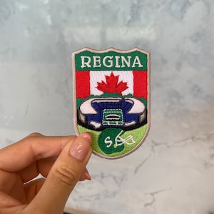 May include: A green, white, and red embroidered patch with the word "REGINA" in white lettering. The patch features a Canadian flag, a stadium, and a hockey helmet.