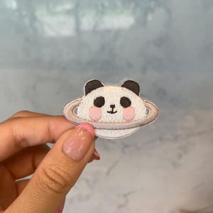 May include: A white panda bear with black ears and eyes is embroidered on a white planet with a silver ring. The planet is surrounded by a pink halo.