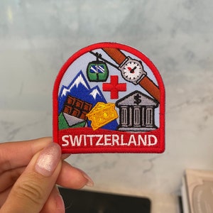 Könnte beinhalten: Ein gestickter Aufnäher in Rot, Weiß und Blau mit der Aufschrift "SWITZERLAND" in weißen Buchstaben. Der Aufnäher zeigt eine Schweizer Flagge, eine Bergkette, eine Schokoladentafel, einen Käseblock, ein Bankgebäude und eine Uhr.