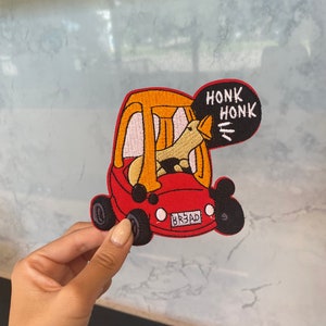 Peut inclure: Écusson brodé représentant un canard conduisant une voiture rouge. Le canard klaxonne, avec les mots "HONK HONK" dans une bulle. La voiture a des roues noires et la plaque d'immatriculation indique "BR3AD".