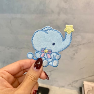 May include: A light blue embroidered elephant patch with a fluffy texture, holding a yellow star. The elephant has a pink and yellow bow. The patch is a cute, whimsical design.