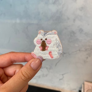 May include: Embroidered patch featuring a cartoon koala bear in white, gray, and pink. The koala has pink cheeks, a brown nose, and is sitting down. The patch is likely for clothing or accessories.