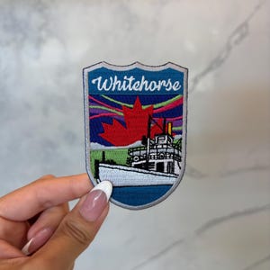 Könnte beinhalten: Gestickter Aufnäher mit dem Schriftzug "Whitehorse", einem roten Ahornblatt und einem Boot. Der Aufnäher hat einen blauen Rand und ein farbenfrohes Aurora Borealis-Design. Der Aufnäher ist schildförmig.
