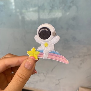May include: Embroidered patch showing a white astronaut with a black visor, sitting on a rainbow arc. A yellow star is next to the astronaut. The patch is designed for application on clothing or accessories.