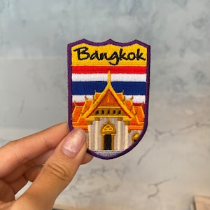 Puede incluir: Un parche bordado de color morado, amarillo y naranja con la palabra "Bangkok" y una representación de un templo tailandés.