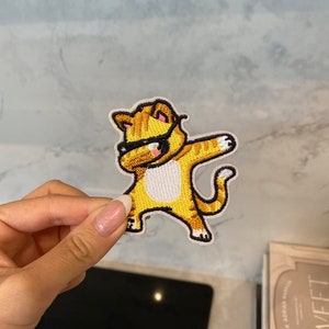 以下が含まれることがあります： 黄色とオレンジ色の刺繍が施されたアイロン接着パッチ。サングラスをかけてダブダンスをしている猫のイラストです。