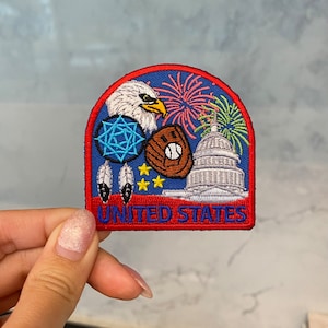 Könnte beinhalten: Ein gestickter Patch in Rot, Weiß und Blau mit einem Adler, Feuerwerk, einem Baseballhandschuh, einem Baseball, einem Traumfänger und dem Kapitol. Der Text "UNITED STATES" befindet sich am unteren Rand des Patches.