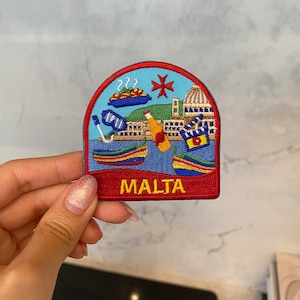 Może przedstawiać: Naszywka haftowana w kolorze czerwonym, białym i niebieskim z przedstawieniem krzyża maltańskiego, budynku, łodzi i napisem "MALTA" w żółtych literach.