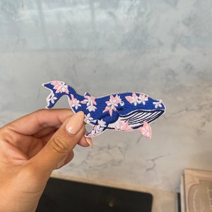 May include: A blue whale embroidered patch with pink flowers. The whale is facing right and has a white outline.