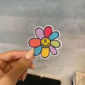 May include: A colourful embroidered flower patch with a yellow smiley face in the centre. The flower has six petals in shades of red, orange, yellow, green, blue, and purple.
