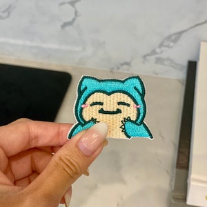 Könnte beinhalten: Ein blauer und grüner gestickter Bügelaufkleber mit einer Cartoonfigur mit einem lächelnden Gesicht. Die Figur trägt ein blaues Kapuzenkleid und hat rosa Wangen.
