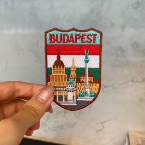 May include: A brown, embroidered patch with a shield shape. The patch features the word "BUDAPEST" in white text on a light blue background. The shield also features a depiction of the Hungarian flag and a cityscape with buildings.