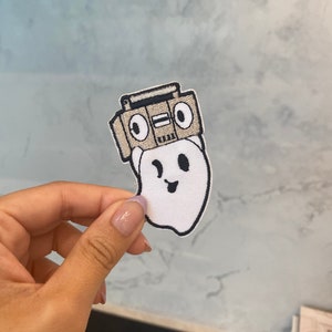 May include: A white ghost with a black smiling face wearing a brown and black boombox on its head. The ghost is embroidered on a white fabric.