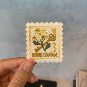 Puede incluir: Un parche blanco bordado con una flor amarilla, dos llaves y el texto "Bonne Courage" en amarillo.