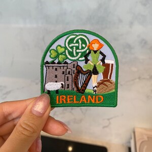 May include: Green and orange embroidered patch with a Celtic knot, shamrock, castle, harp, and a woman in a green dress. The text "IRELAND" is on the bottom of the patch.