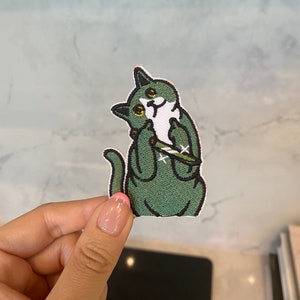 May include: Green embroidered iron-on patch of a cat holding a knife with a white face and yellow eyes.