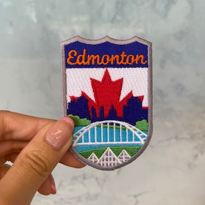 Può includere: Toppa ricamata con il nome "Edmonton" in arancione. Il design include una foglia d'acero rossa, un ponte e uno skyline su sfondo blu. La toppa ha un bordo grigio.