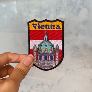May include: A red, white, and yellow embroidered patch with the word "Vienna" in blue lettering. The patch features a stylized image of a building with a green dome.