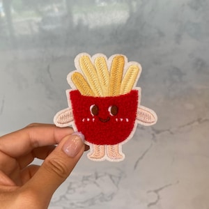 Puede incluir: Un parche de fieltro rojo y amarillo de un personaje de dibujos animados de papas fritas con una cara sonriente, brazos y piernas.