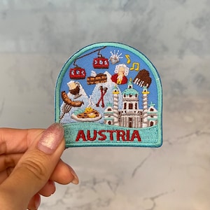 May include: A blue and green embroidered patch with a white background featuring various Austrian landmarks and symbols, including a mountain range, a building, a musical note, a person, and a gondola. The text "AUSTRIA" is embroidered in red at the bottom of the patch.