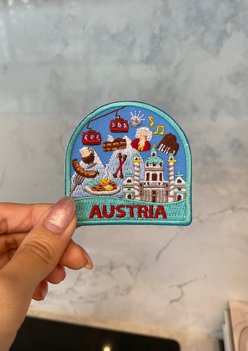 May include: A blue and green embroidered patch with a white background featuring various Austrian landmarks and symbols, including a mountain range, a building, a musical note, a person, and a gondola. The text "AUSTRIA" is embroidered in red at the bottom of the patch.