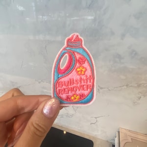 May include: An embroidered iron-on patch shaped like a bottle of detergent with the text "Bullshit Remover" on the front. The patch is pink and blue.