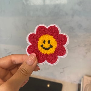 Puede incluir: Parche de fieltro rojo en forma de flor con una carita sonriente amarilla en el centro. La flor tiene costuras blancas alrededor de los bordes.