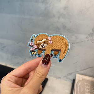 May include: Embroidered patch featuring a cartoon sloth with brown fur, a light blue outline, and a sprig of pink flowers. The sloth is lying down, and the patch is approximately 3 inches wide.