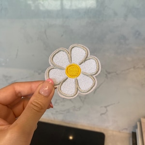 Może przedstawiać: Biała haftowana łatka z motywem stokrotki z żółtym środkiem.