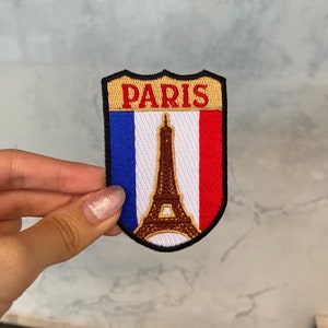 May include: A brown and gold embroidered patch with the word "PARIS" in red lettering. The patch features a white background with a blue and red stripe on either side, and a brown outline of the Eiffel Tower in the center.