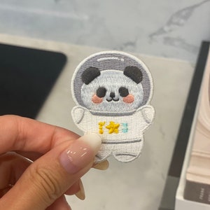 May include: An embroidered patch featuring a panda wearing an astronaut suit, with a yellow star on the chest. The panda has black ears and eyes and pink cheeks.