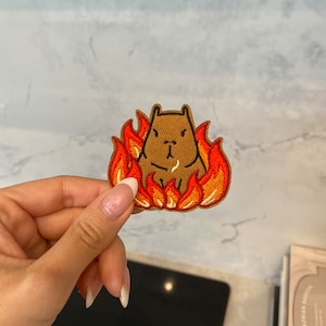 Peut inclure: Patch brodé représentant un capybara brun avec une expression renfrognée, entouré de flammes orange.