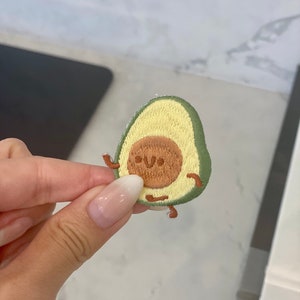 May include: A green and brown embroidered avocado patch with a smiling face and small legs.