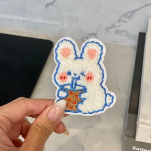Könnte beinhalten: Gestickter Aufnäher mit einem weißen Hasen mit blauem Umriss, rosa Wangen und Ohren, der aus einer braunen Tasse mit blauem Strohhalm trinkt. Der Aufnäher hat eine flauschige Textur, für Kleidung oder Accessoires.