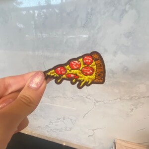 Puede incluir: Parche de una porción de pizza con pepperoni, queso y corteza marrón. El queso amarillo gotea por el costado. El parche se muestra sobre un fondo blanco y gris.