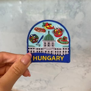 May include: A blue and white embroidered patch with a building, a Rubik's cube, a pen, and various food items. The text "HUNGARY" is embroidered in yellow on the bottom of the patch.