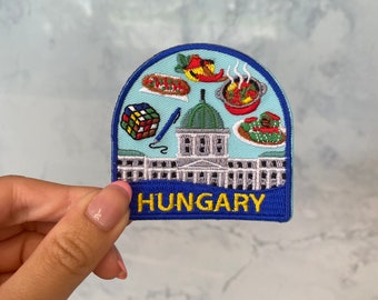 May include: A blue and white embroidered patch with a building, a Rubik's cube, a pen, and various food items. The text "HUNGARY" is embroidered in yellow on the bottom of the patch.
