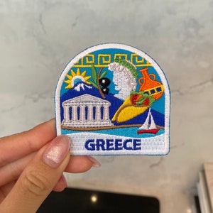 Könnte beinhalten: Ein blauer und weißer gestickter Aufnäher mit einer Darstellung eines griechischen Tempels, Olivenzweigen, einer Sonne, einem Krug und einem Segelboot. Der Text "GREECE" ist in Weiß auf der Unterseite des Aufnähers gestickt.