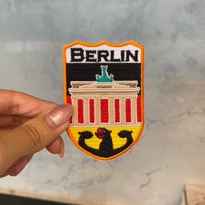 Puede incluir: Un parche bordado amarillo, negro y rojo con la palabra "BERLIN" en letras negras. El parche presenta una imagen estilizada de la Puerta de Brandeburgo y un pájaro negro con un pico rojo.