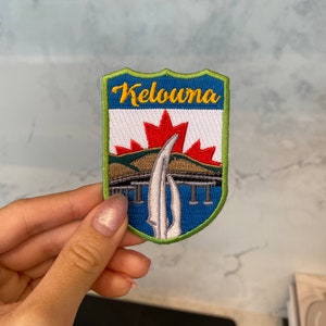 Peut inclure: Écusson brodé avec un fond bleu et une bordure verte. L'écusson présente une feuille d'érable blanche avec un centre rouge, un pont et le texte "Kelowna" en jaune.