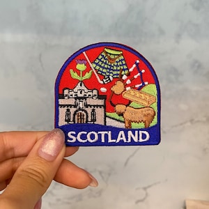 Könnte beinhalten: Ein gestickter Aufnäher in Rot, Blau und Weiß mit einem Schloss, einer Distel, einem Kilt, Dudelsäcken und einem Schaf. Der Text "SCOTLAND" ist in Weiß auf blauem Grund gestickt.