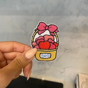 May include: An embroidered iron-on patch depicting a yellow basket overflowing with pink and red hearts. A pink bow sits atop the basket. The basket has a blue label with the word "Love" on it.
