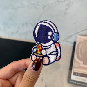 May include: An embroidered patch featuring an astronaut in a white spacesuit, wearing a blue helmet and holding a slice of pizza. The astronaut is sitting down with pink legs.