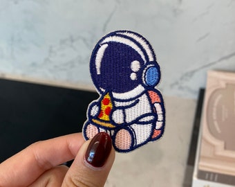May include: A white embroidered patch of an astronaut wearing a blue helmet and holding a slice of pizza. The astronaut is sitting down with pink legs and a white body.