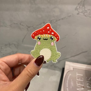 May include: An embroidered green frog wearing a red and white mushroom hat. The frog has a white belly and pink cheeks.