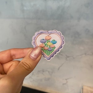 Op de afbeelding: Een geborduurde patch in de vorm van een hart met een bloemenmotief. De bloemen zijn in tinten roze, blauw, geel en groen. De patch heeft een gegolfde rand en is omgeven door een paarse omtrek.