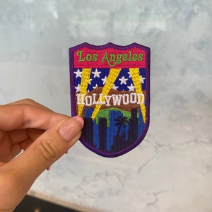 May include: Purple, blue, green, yellow and red embroidered patch with the text "Los Angeles" and "HOLLYWOOD" on a white background. The patch features a cityscape with palm trees and spotlights.